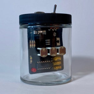Peut inclure: Un bocal en verre transparent avec un couvercle noir contenant un petit circuit imprimé avec divers composants électroniques, notamment des résistances, des condensateurs et une LED rouge.