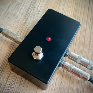 May include: A black effects pedal with a silver footswitch and a red LED indicator. Two black cables with silver connectors are plugged into the side. The pedal is on a wooden surface.