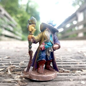 May include: A miniature wizard figurine with a long white beard, wearing a purple cloak and a wide-brimmed hat. He holds a staff with a detailed top. The wizard's outfit includes a blue shirt, brown boots, and a gold-colored coat. The figurine stands on a brown base.