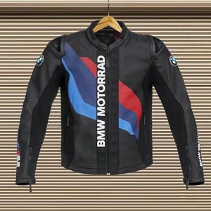 May include: Black motorcycle jacket with the text "BMW MOTORRAD" in white. The jacket has blue and red accents and BMW logos on the sleeves. It is hanging on a wooden hanger.