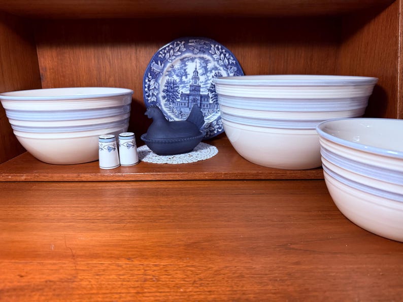 Op de afbeelding: Een verzameling keramische kommen met blauw-witte strepen, een blauw-wit decoratief bord, een zwart kipvormig bakje en twee kleine zout- en pepervaatjes. De items zijn gerangschikt op een houten plank.