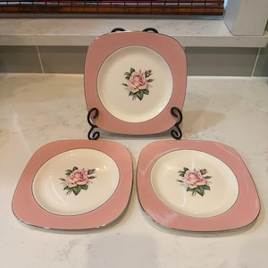 May include: Set of three square-shaped pink plates with a floral rose design in the center. The plates have a silver trim and are displayed on a black metal stand. The plates are a light pink color.