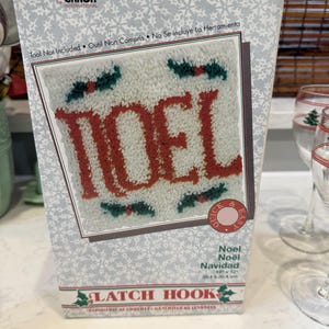 May include: A WonderArt by Caron International latch hook kit box featuring a white background with a snowflake pattern. The box displays a finished latch hook project with the red text NOEL surrounded by green holly leaves, measuring 12 x 12 inches.