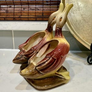 May include: A pair of vintage ceramic duck planters. The ducks are in a kissing pose, with yellow, brown, and burgundy coloring. The planters have open compartments for plants or decor. The base is a square shape.