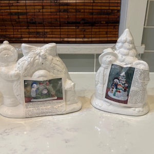 May include: Two white ceramic Christmas decorations. One features a snowman holding a wreath, the other a snowman next to a Christmas tree. Each has a colorful image of snowmen and a Christmas tree.
