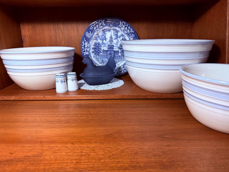 Op de afbeelding: Een verzameling keramische kommen met blauwe en beige strepen, een blauw-wit decoratief bord, een zwart kippenvormig bakje en twee zout- en pepervaatjes. De items worden tentoongesteld op een houten plank.