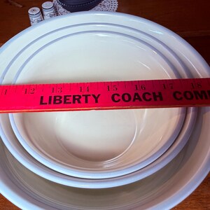Op de afbeelding: Drie in elkaar geneste witte keramische kommen met lichtblauwe randen en een dunne blauwe streep. Een rode liniaal met de tekst "LIBERTY COACH COMPANY" ligt over de kommen. De kommen staan op een houten oppervlak.