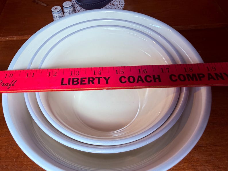 Op de afbeelding: Drie in elkaar geneste witte keramische kommen met lichtblauwe randen en een dunne blauwe streep. Een rode liniaal met de tekst "LIBERTY COACH COMPANY" ligt over de kommen. De kommen staan op een houten oppervlak.