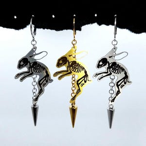 May include: Three dangling earrings featuring a cutout skeleton rabbit design. The left and right earrings are silver-toned, while the center earring is gold-toned. Each rabbit figure is attached to a small chain ending in a pointed metal spike charm.