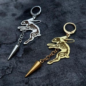 May include: Two hoop earrings featuring a rabbit skeleton charm attached to a small chain and a pointed cone-shaped pendant. One earring is silver-toned metal, and the other is gold-toned metal, both displaying detailed skeletal anatomy.