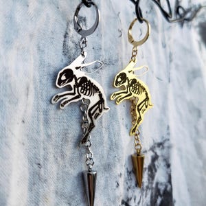 May include: Two dangling earrings featuring a silver-tone and a gold-tone metal rabbit skeleton charm. Each earring hangs from a circular hoop and includes a short chain ending in a pointed, cone-shaped metal spike charm suspended below the rabbit.