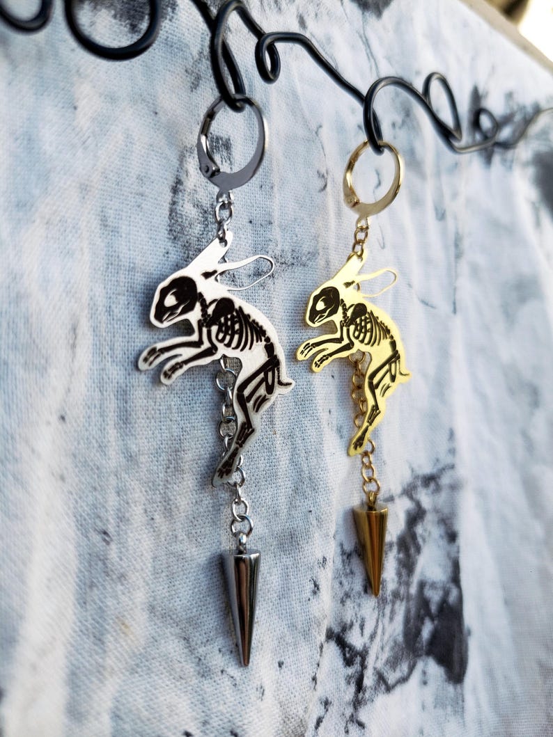 May include: Two dangling earrings featuring a silver-tone and a gold-tone metal rabbit skeleton charm. Each earring hangs from a circular hoop and includes a short chain ending in a pointed, cone-shaped metal spike charm suspended below the rabbit.