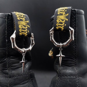 Puede incluir: Un par de botas de cuero negro con tiradores de talón negros con texto amarillo AirWair. Cada tirador está adornado con un anillo hexagonal segmentado de tono plateado que sostiene un dije de metal colgante en forma de estrella de cuatro puntas.
