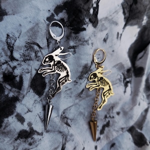 May include: Two dangling earrings featuring a silver-tone and a gold-tone rabbit skeleton charm. Each earring includes a circular hoop, a chain segment, and a pointed cone-shaped spike charm hanging at the bottom, showcasing a detailed skeletal design.