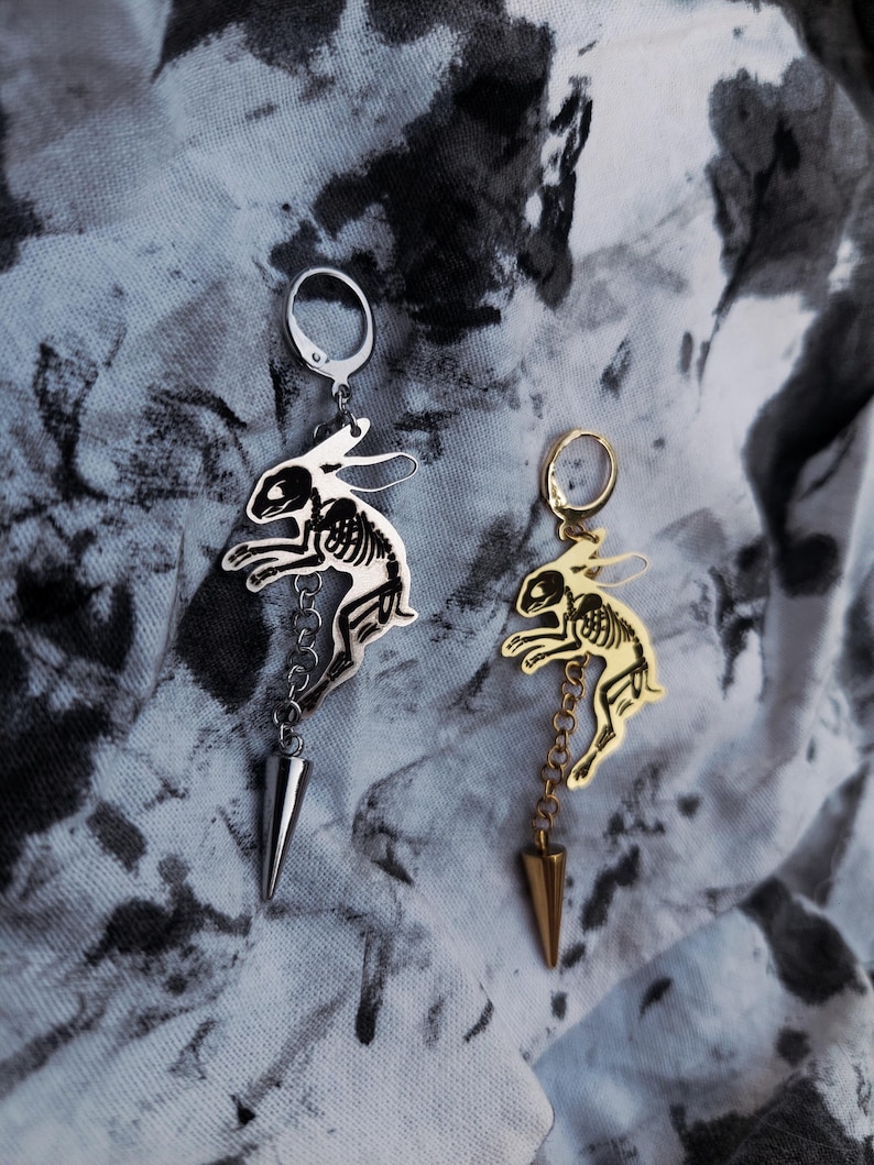 May include: Two dangling earrings featuring a silver-tone and a gold-tone rabbit skeleton charm. Each earring includes a circular hoop, a chain segment, and a pointed cone-shaped spike charm hanging at the bottom, showcasing a detailed skeletal design.
