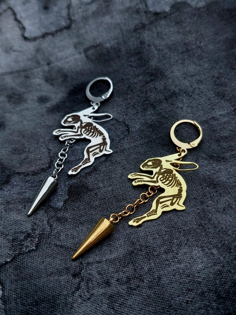 May include: Two hoop earrings featuring a rabbit skeleton charm attached to a small chain and a pointed cone-shaped pendant. One earring is silver-toned metal, and the other is gold-toned metal, both displaying detailed skeletal anatomy.
