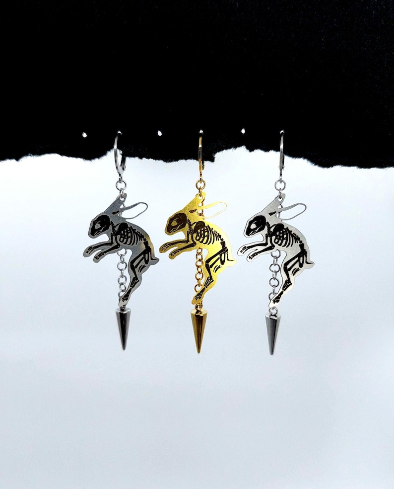 May include: Three dangling earrings featuring a cutout skeleton rabbit design. The left and right earrings are silver-toned, while the center earring is gold-toned. Each rabbit figure is attached to a small chain ending in a pointed metal spike charm.