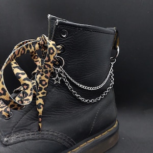Könnte beinhalten: Ein schwarzer Lederstiefel mit Schnürsenkeln im Leopardenmuster und einem silberfarbenen Metallketten-Accessoire an den Ösen. Die Kette enthält einen kleinen sternförmigen Anhänger, und der Stiefel hat eine dicke Sohle mit sichtbaren gelben Nähten.