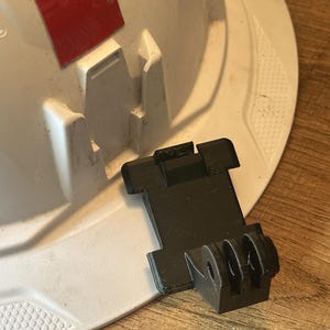 Könnte beinhalten: Eine schwarze Kunststoff-Halterung, die an der Vorderseite eines weißen Schutzhelms befestigt ist. Die Halterung verfügt über ein dreizinkiges Stecksystem, das häufig für Action-Kameras verwendet wird, und sitzt fest im geformten Schlitz am Helmrand.