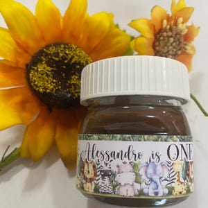 Op de afbeelding: Een klein glazen potje gevuld met een bruine substantie, waarschijnlijk een spread, met een witte deksel. De pot is versierd met een label met de tekst "Alessandro is ONE" en cartoon dieren. Gele zonnebloemen op de achtergrond.