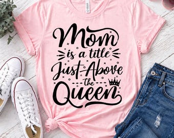 May include: A light pink t-shirt with the text "Mom is a title Just Above the Queen" in black script. The shirt is paired with blue jeans, white sneakers, and sunglasses.