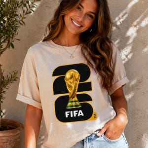May include: Beige t-shirt featuring a graphic of the FIFA World Cup trophy. The design includes the number 23 in black and gold, with the word "FIFA" in white. The shirt is a casual style.