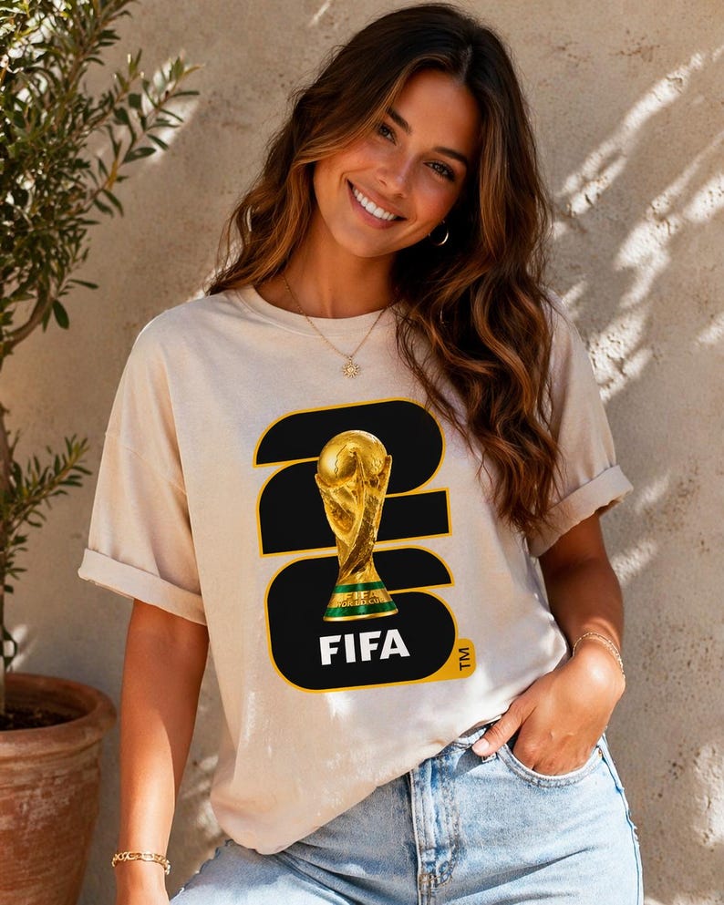 May include: Beige t-shirt featuring a graphic of the FIFA World Cup trophy. The design includes the number 23 in black and gold, with the word "FIFA" in white. The shirt is a casual style.