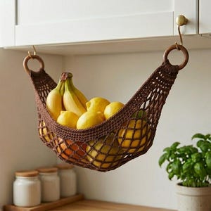 May include: A brown, crocheted fruit hammock suspended from a kitchen cabinet. The hammock is filled with yellow lemons, bananas, and oranges. The kitchen has white cabinets, a window, and a small potted plant.