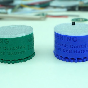 May include: Four cylindrical objects in red, green, blue, and white. Each has a white top and a black protrusion. The sides display the word "WARNING" and text about a cell battery.