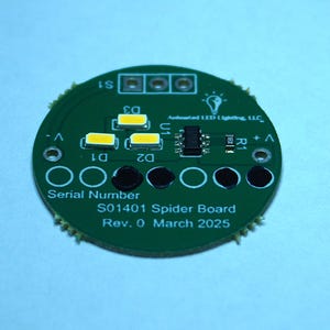 May include: A circular green circuit board with several components, including LED lights and a small integrated circuit. The board has the text "S01401 Spider Board" and "Rev. 0 March 2025" printed on it. The background is a light blue.