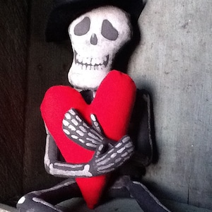 May include: A skeleton doll wearing a black hat holds a red heart. The doll is made of fabric and has a painted skull face. The text "Hexie Hollow Art Dolls 2012" is written below the doll.