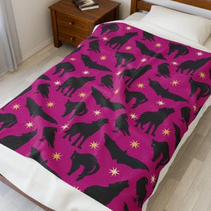 May include: A magenta blanket with a repeating pattern of black wolves, bats, cats, and crows, along with gold starbursts. The blanket is on a bed with a white pillow and a wooden nightstand.