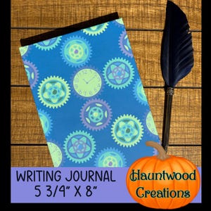 May include: A blue writing journal with a clock and gear design, measuring 14.6 cm x 20.3 cm. A black feather quill and an orange pumpkin are next to the journal. The text "Writing Journal" and "Hauntwood Creations" are visible.