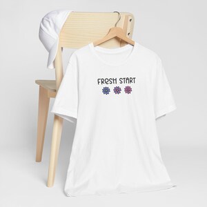 May include: White t-shirt with the text "FRESH START" above three flower graphics. The flowers are blue and pink. A white baseball cap rests on the back of a wooden chair.