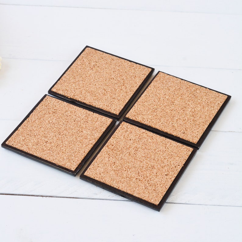 May include: Four square coasters with a cork surface and a dark brown border. The coasters are arranged in a diamond shape on a white wooden surface. Each coaster is approximately 4 inches square.