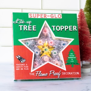 May include: A vintage Christmas tree topper in a star shape, encased in a box with green, red, and white accents. The box reads "SUPER-GLO Lite-up TREE TOPPER" and "THE Flame Proof DECORATION". The star has a silver tinsel appearance with colorful lights.