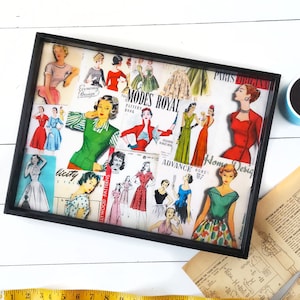 May include: A black tray with a white background filled with vintage sewing patterns. The patterns feature women's clothing designs with various colors and styles. The tray has a black frame and is filled with a variety of patterns, including "Modes Royal", "Advance 6085", and "Home Design".