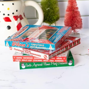 May include: A stack of vintage Christmas decoration boxes. The boxes are red, green, and blue with text that reads "Wilson's Christmas Decorations" and "Christmas Tree Ornaments". The boxes are stacked in front of a snowman pitcher and miniature Christmas trees.