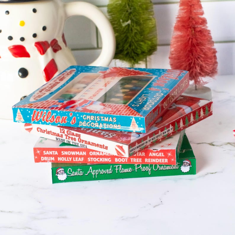 May include: A stack of vintage Christmas decoration boxes. The boxes are red, green, and blue with text that reads "Wilson's Christmas Decorations" and "Christmas Tree Ornaments". The boxes are stacked in front of a snowman pitcher and miniature Christmas trees.