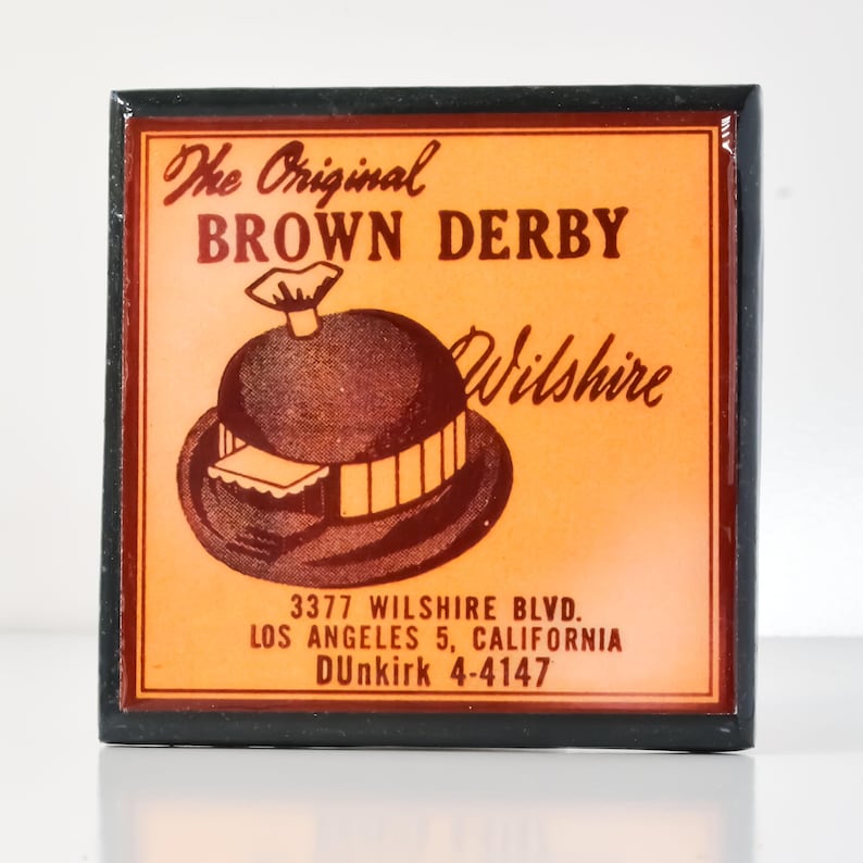 May include: A vintage sign for the "The Original Brown Derby Wilshire" restaurant in Los Angeles, California. The sign features a brown derby hat with a white brim and a red and white striped awning. The sign also includes the address "3377 Wilshire Blvd. Los Angeles 5, California" and the phone number "Dunkirk 4-4147".