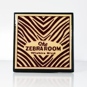 May include: A black and white zebra print square sign with the text "The Zebra Room, Wilshire Blvd" in red.