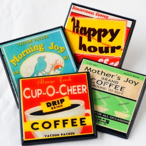 Op de afbeelding: Vier vintage-stijl koffieonderzetters met zwarte frames. De onderzetters hebben kleurrijke ontwerpen en tekst, waaronder "Morning Joy", "Cup-O-Cheer", "Happy Hour" en "Mother's Joy Brand Coffee".