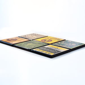 May include: Six square coasters with black frames and vintage-style designs. The coasters feature images of a hat, a plant, an ice cream cone, a building, a mountain, and a restaurant.