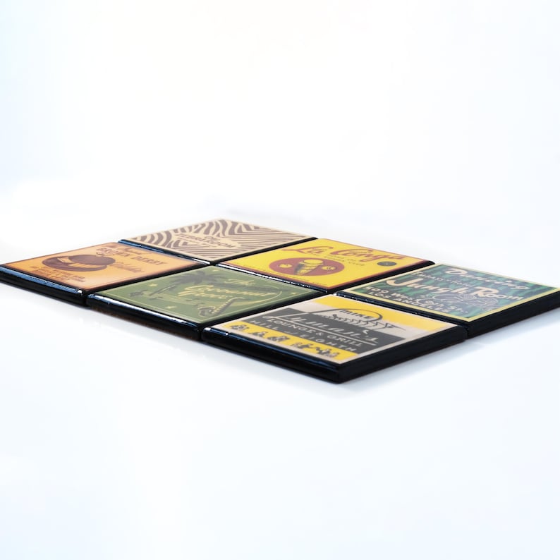 May include: Six square coasters with black frames and vintage-style designs. The coasters feature images of a hat, a plant, an ice cream cone, a building, a mountain, and a restaurant.