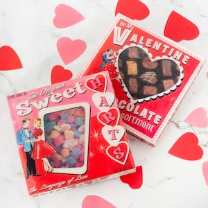 May include: Two vintage Valentine's Day candy boxes. One is pink and red with "Sweet Hearts" and a window showing heart-shaped candies. The other is red with a heart-shaped window displaying chocolates and the words "Be My Valentine".