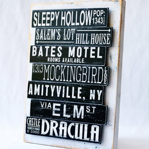 Pu&ograve; includere: Un cartello di legno bianco con testo dipinto in nero che elenca luoghi famosi di film horror. Il testo include Sleepy Hollow, Salem's Lot, Bates Motel, 1313 Mockingbird Lane, Amityville, NY, via Elm St e Castle Dracula.