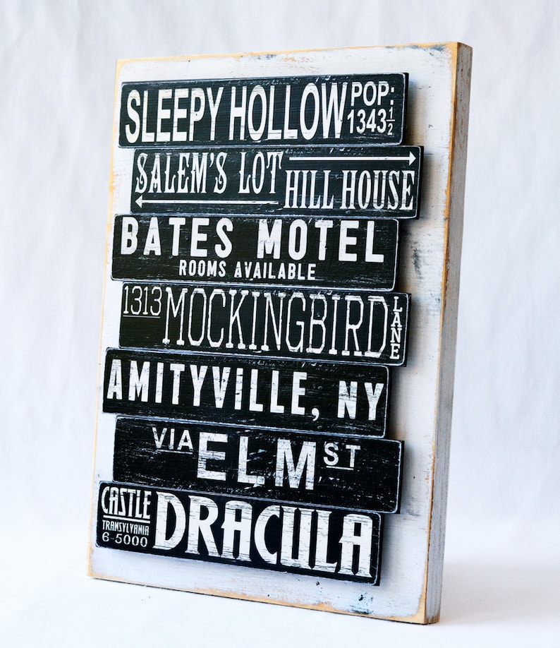 Pu&ograve; includere: Un cartello di legno bianco con testo dipinto in nero che elenca luoghi famosi di film horror. Il testo include Sleepy Hollow, Salem's Lot, Bates Motel, 1313 Mockingbird Lane, Amityville, NY, via Elm St e Castle Dracula.