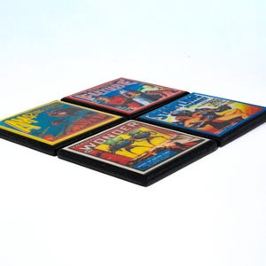 May include: Four square coasters with vintage comic book covers. The covers feature illustrations of a man in a red suit, a man in a blue suit, a man in a green suit, and a man in a yellow suit. The coasters are black with a wood grain finish.