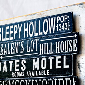 Pu&ograve; includere: Un cartello di legno con testo dipinto in bianco e nero. Il cartello recita "Sleepy Hollow Pop: 1343 1/2", "Salem's Lot Hill House", "Bates Motel Camere disponibili".