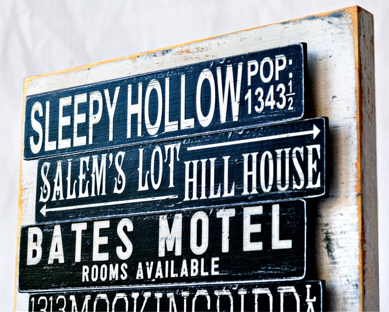 Pu&ograve; includere: Un cartello di legno con testo dipinto in bianco e nero. Il cartello recita "Sleepy Hollow Pop: 1343 1/2", "Salem's Lot Hill House", "Bates Motel Camere disponibili".